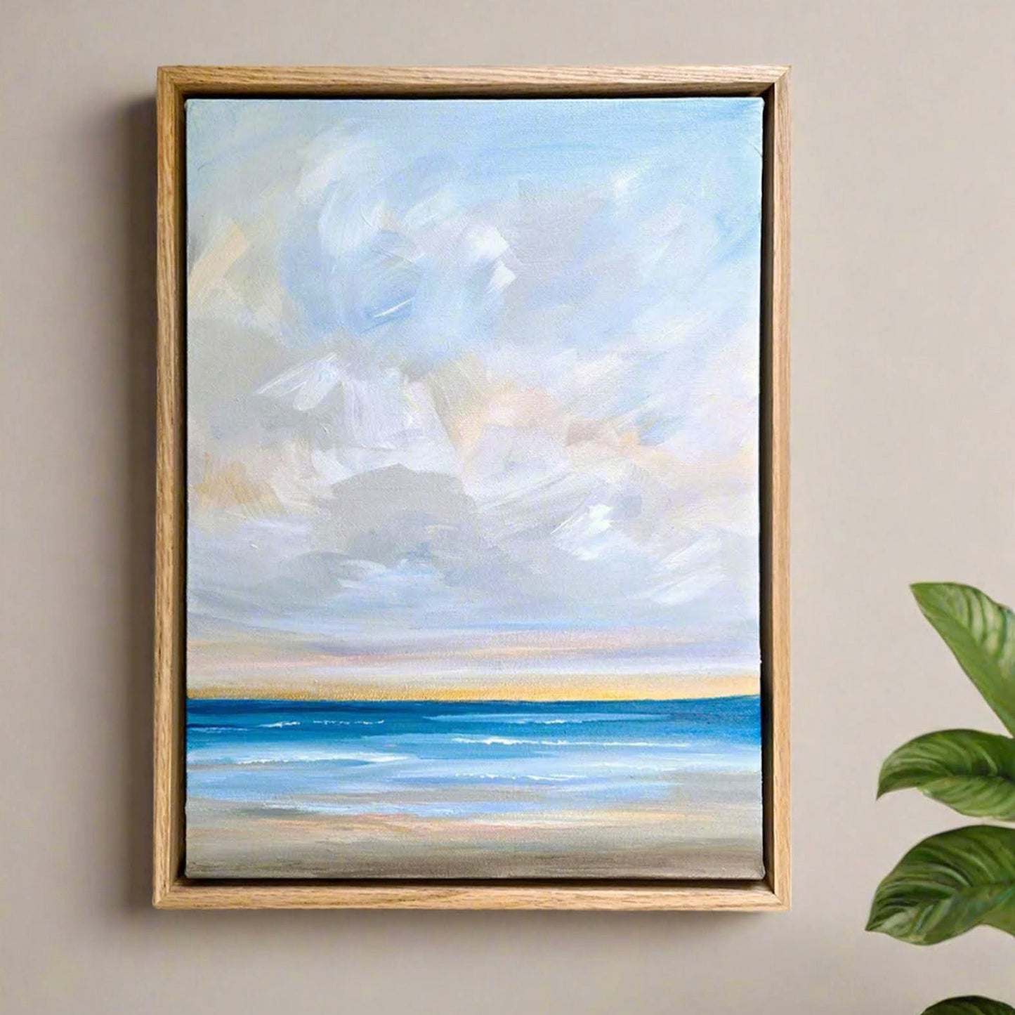Framed painting of a beach scene with blue sky and water on a beige wall.