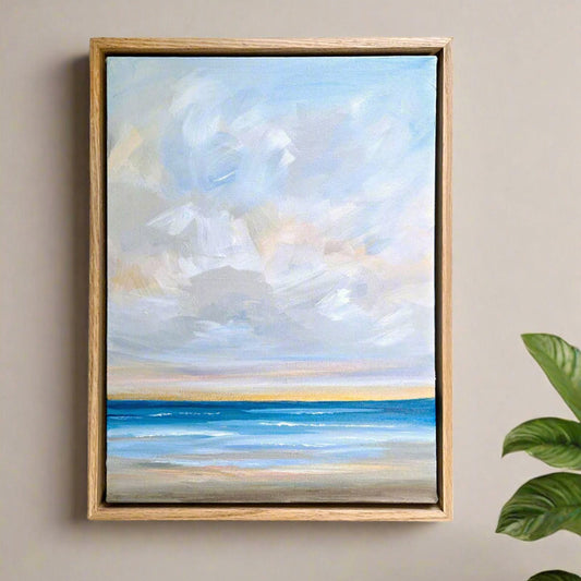 Framed painting of a beach scene with blue sky and water on a beige wall.